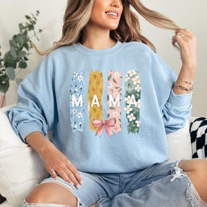 Pu&ograve; includere: Felpa blu chiaro con un design floreale e la parola "MAMA" in lettere bianche. Il design floreale &egrave; in tonalit&agrave; di rosa, giallo e verde.