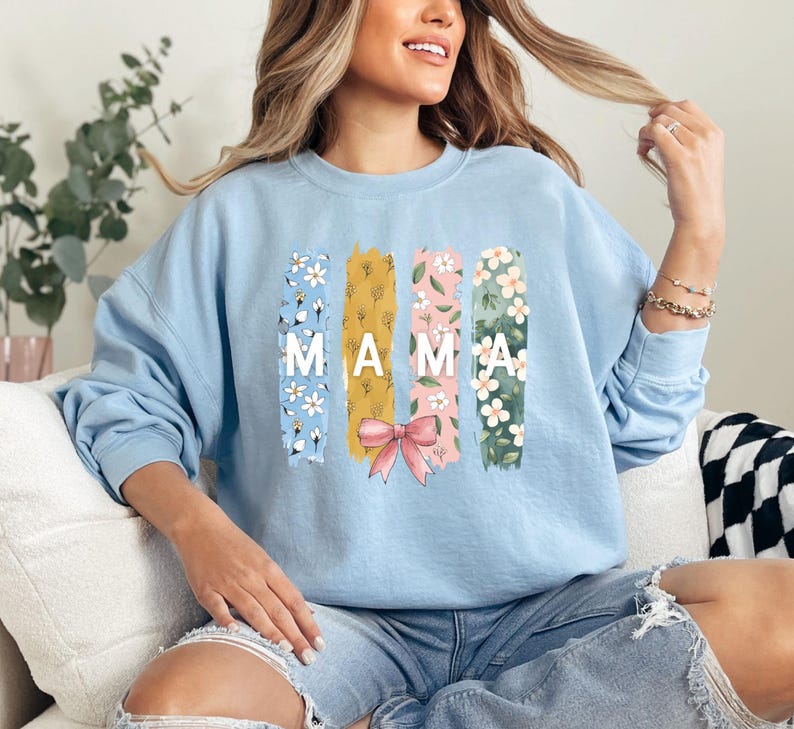 Pu&ograve; includere: Felpa blu chiaro con un design floreale e la parola "MAMA" in lettere bianche. Il design floreale &egrave; in tonalit&agrave; di rosa, giallo e verde.