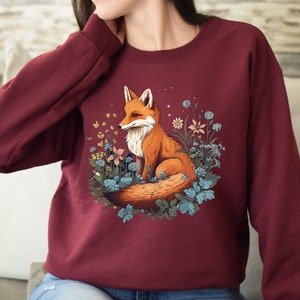May include: A maroon long-sleeve sweatshirt featuring a graphic print of an orange fox sitting among blue and yellow flowers. The fox has a white chest and a bushy tail, surrounded by various botanical elements and small white star-like accents.