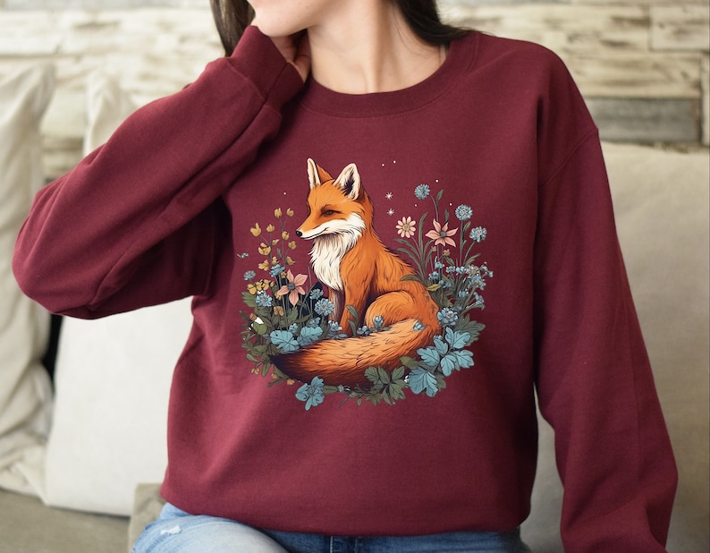 May include: A maroon long-sleeve sweatshirt featuring a graphic print of an orange fox sitting among blue and yellow flowers. The fox has a white chest and a bushy tail, surrounded by various botanical elements and small white star-like accents.