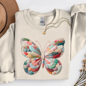 May include: Cream-colored sweatshirt featuring a colorful butterfly design. The butterfly's wings are composed of various fabric patches in shades of red, blue, green, and yellow. A gold necklace with a star pendant and a brown hat are also visible.