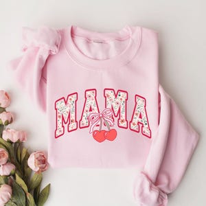 Może przedstawiać: R&oacute;żowa bluza z nadrukiem "MAMA" w kwiatowym wzorze i dwiema czerwonymi wiśniami z r&oacute;żową kokardką.