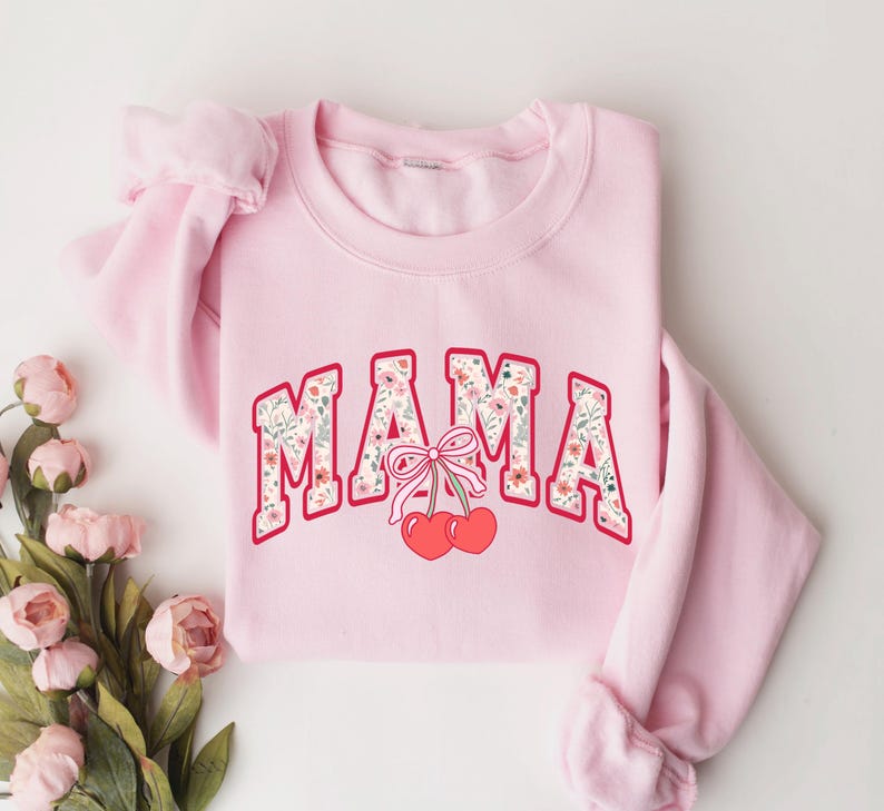 Może przedstawiać: R&oacute;żowa bluza z nadrukiem "MAMA" w kwiatowym wzorze i dwiema czerwonymi wiśniami z r&oacute;żową kokardką.
