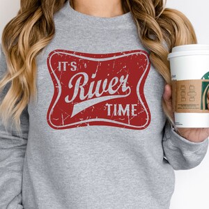 May include: A heather gray long-sleeve sweatshirt featuring a distressed red rectangular graphic with rounded corners. The graphic contains the white text "IT'S River TIME" in a stylized font, mimicking a vintage beverage label design.