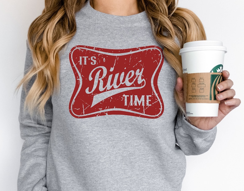 May include: A heather gray long-sleeve sweatshirt featuring a distressed red rectangular graphic with rounded corners. The graphic contains the white text "IT'S River TIME" in a stylized font, mimicking a vintage beverage label design.