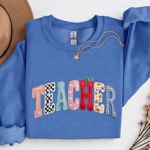 May include: A blue sweatshirt with the word "TEACHER" embroidered in colourful, patterned letters. The letters feature various designs, including checkered, polka dot, and rainbow patterns. A gold necklace with a star pendant is draped across the sweatshirt.
