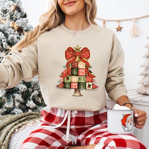 Puede incluir: Una sudadera beige con un diseño de árbol de Navidad con un lazo rojo y cuadrados de patchwork en rojo, verde y dorado. La persona lleva unos pantalones cortos de cuadros rojos y blancos y sostiene una taza con un gráfico de Papá Noel.