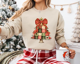 Può includere: Una felpa beige con un design ad albero di Natale con un fiocco rosso e quadrati patchwork rossi, verdi e oro. La persona indossa pantaloncini a quadri rossi e bianchi e tiene in mano una tazza con una grafica di Babbo Natale.