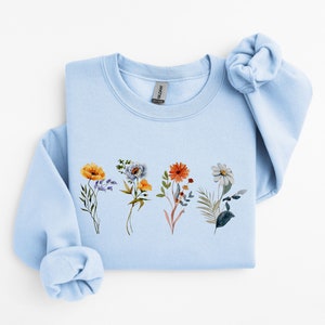 May include: A light blue sweatshirt with a floral print featuring four different types of flowers in shades of yellow, orange, blue, and white.