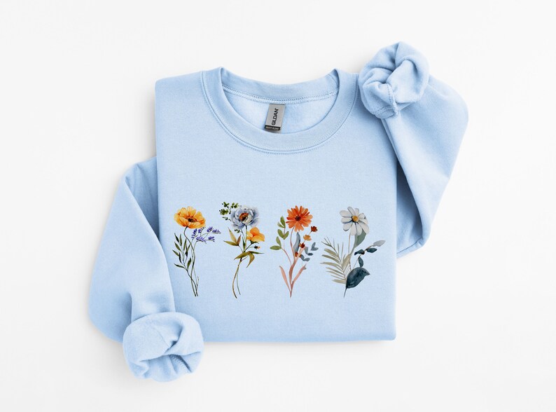 May include: A light blue sweatshirt with a floral print featuring four different types of flowers in shades of yellow, orange, blue, and white.