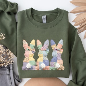 May include: Olive green sweatshirt featuring three patchwork bunny designs with pastel-colored ears and bowties. The bunnies have a variety of patterned fabrics, including floral and polka dots. The sweatshirt is folded, with a pair of jeans and dried flowers in the background.