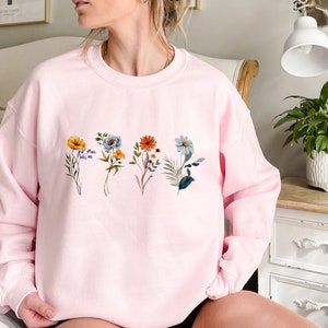 May include: A pink sweatshirt with a floral design featuring five different flowers in shades of blue, orange, yellow, and green.