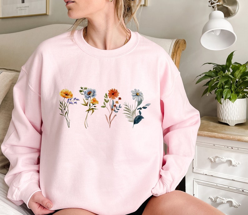 May include: A pink sweatshirt with a floral design featuring five different flowers in shades of blue, orange, yellow, and green.