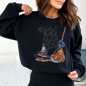 May include: A black long-sleeve sweatshirt featuring a graphic print of a blue witch hat with autumn leaves and a wooden broomstick. The text "Yes, I can drive a stick!" is printed in a stylized black script font above the hat and broomstick.