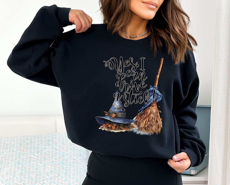 May include: A black long-sleeve sweatshirt featuring a graphic print of a blue witch hat with autumn leaves and a wooden broomstick. The text "Yes, I can drive a stick!" is printed in a stylized black script font above the hat and broomstick.
