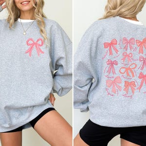 May include: A heather gray crewneck sweatshirt featuring a small pink bow graphic on the front left chest. The back displays a large collection of various pink bows arranged in a grid, accompanied by cursive text including words like Loved, Brave, and Precious.