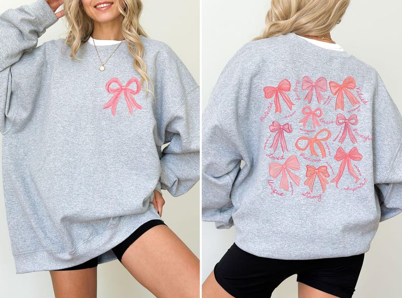 May include: A heather gray crewneck sweatshirt featuring a small pink bow graphic on the front left chest. The back displays a large collection of various pink bows arranged in a grid, accompanied by cursive text including words like Loved, Brave, and Precious.