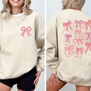 May include: A beige crewneck sweatshirt featuring a small pink bow graphic on the front left chest. The back displays a large collection of various pink bows accompanied by cursive text including words like Loved, Precious, Strong, and Unique.