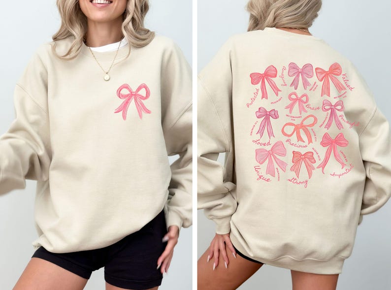 May include: A beige crewneck sweatshirt featuring a small pink bow graphic on the front left chest. The back displays a large collection of various pink bows accompanied by cursive text including words like Loved, Precious, Strong, and Unique.