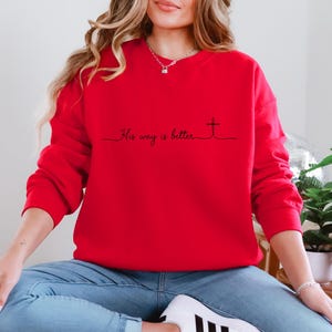 May include: A bright red sweatshirt with the phrase "His way is better" and a cross design in black. The long-sleeved top is made of a soft material and is suitable for casual wear.