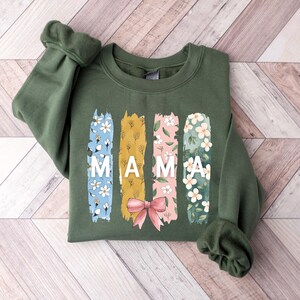 Pu&ograve; includere: Una felpa verde con un design floreale e la parola "MAMA" stampata sul davanti.