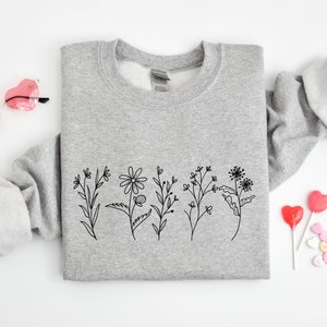 May include: A heather gray crewneck sweatshirt featuring a black line art illustration of five distinct wildflowers across the center. The garment has long sleeves with ribbed cuffs and a ribbed neckline, presented with heart-shaped glasses and candy.