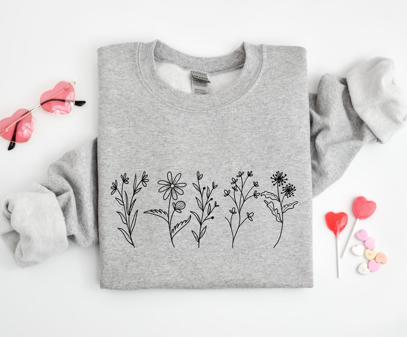 May include: A heather gray crewneck sweatshirt featuring a black line art illustration of five distinct wildflowers across the center. The garment has long sleeves with ribbed cuffs and a ribbed neckline, presented with heart-shaped glasses and candy.