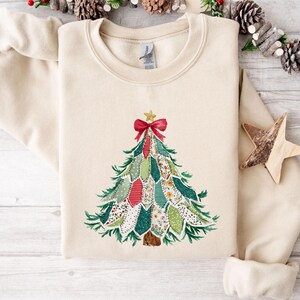 Puede incluir: Sudadera de color crema con un diseño bordado de árbol de Navidad. El árbol está compuesto por parches de tela en verde, rojo y estampados florales, coronado con un lazo rojo y una estrella dorada. Una prenda navideña acogedora.