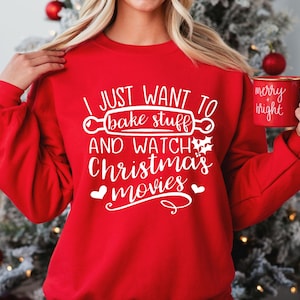 May include: A red long-sleeve sweatshirt featuring white text that reads I JUST WANT TO BAKE STUFF AND WATCH Christmas movies with a rolling pin graphic and holly leaves. A matching red mug with the text merry + bright is held in one hand.
