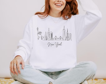 Könnte beinhalten: Weißes Sweatshirt mit einer schwarzen Strichzeichnung der Skyline von New York City, einschließlich der Freiheitsstatue, Wolkenkratzern und der Brooklyn Bridge. Das Wort "New York" steht darunter in Schreibschrift.