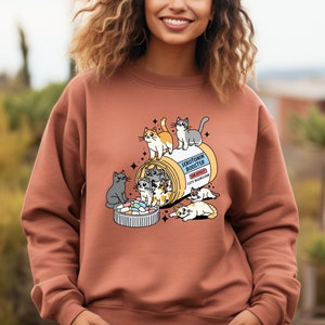 K&ouml;nnte beinhalten: Ein terrakottafarbenes Sweatshirt mit Rundhalsausschnitt und einer Grafik einer Pillendose mit der Aufschrift Serotonin Booster sowie dem Text Side Effect Cute Aggression. Das Motiv zeigt mehrere illustrierte Katzen, die mit der Dose und bunten Pillen spielen.