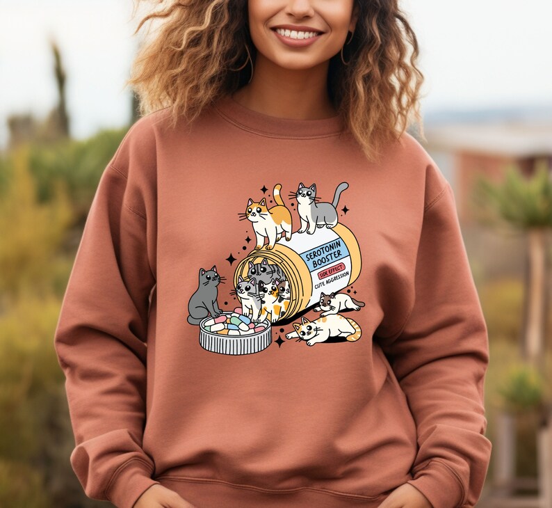 K&ouml;nnte beinhalten: Ein terrakottafarbenes Sweatshirt mit Rundhalsausschnitt und einer Grafik einer Pillendose mit der Aufschrift Serotonin Booster sowie dem Text Side Effect Cute Aggression. Das Motiv zeigt mehrere illustrierte Katzen, die mit der Dose und bunten Pillen spielen.