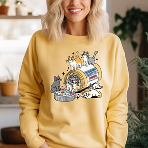 K&ouml;nnte beinhalten: Ein gelbes Sweatshirt mit Rundhalsausschnitt und einer Grafik einer Pillendose mit der Aufschrift Serotonin Booster sowie dem Text Side Effect Cute Aggression. Mehrere gezeichnete Katzen sind um und in der Dose sowie einem Deckel voller Pillen zu sehen.