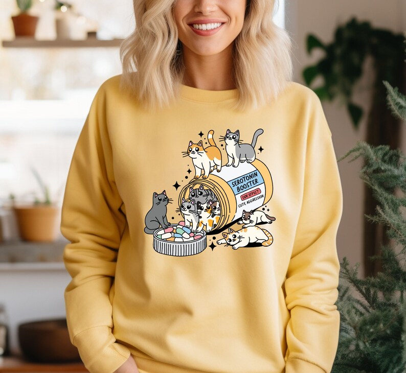K&ouml;nnte beinhalten: Ein gelbes Sweatshirt mit Rundhalsausschnitt und einer Grafik einer Pillendose mit der Aufschrift Serotonin Booster sowie dem Text Side Effect Cute Aggression. Mehrere gezeichnete Katzen sind um und in der Dose sowie einem Deckel voller Pillen zu sehen.