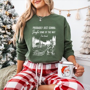 Puede incluir: Sudadera verde con el texto "Probably Just Gonna Jingle Some of the Way I'm Tired". El gr&aacute;fico muestra a una persona tirando de un trineo con regalos a trav&eacute;s de un pueblo nevado. Una taza de Pap&aacute; Noel se sostiene en la mano.