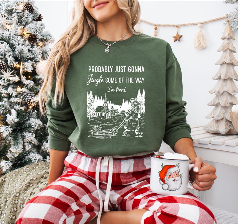 Puede incluir: Sudadera verde con el texto "Probably Just Gonna Jingle Some of the Way I'm Tired". El gr&aacute;fico muestra a una persona tirando de un trineo con regalos a trav&eacute;s de un pueblo nevado. Una taza de Pap&aacute; Noel se sostiene en la mano.