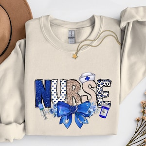 Könnte beinhalten: Cremefarbenes Sweatshirt mit dem Wort "NURSE" in einem dekorativen Design. Die Buchstaben sind in Blau, Leopardenmuster und Braun mit einer blauen Schleife, einer Spritze, einer Medikamentenflasche und einer Krankenschwestermütze.