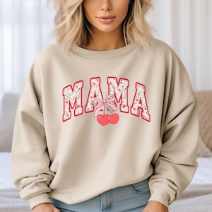 Może przedstawiać: Beżowa bluza z nadrukiem kwiatowym i napisem "MAMA" w kolorze czerwonym z r&oacute;żową kokardką i wiśniami.