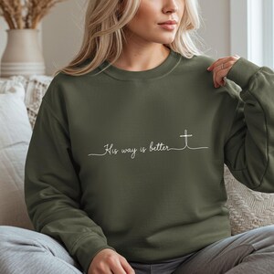 May include: An olive green sweatshirt with the phrase "His way is better" in white script and a cross graphic. The sweatshirt has long sleeves and a crew neck.