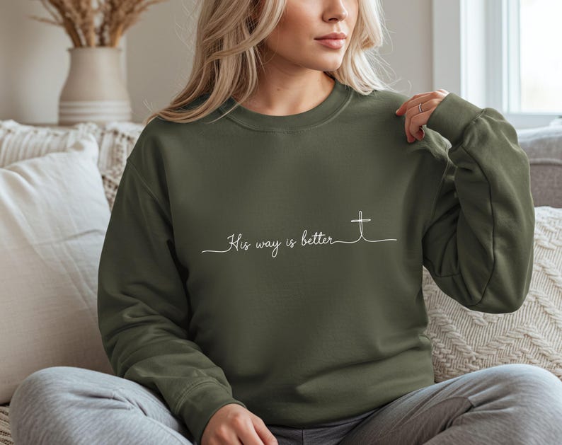 May include: An olive green sweatshirt with the phrase "His way is better" in white script and a cross graphic. The sweatshirt has long sleeves and a crew neck.