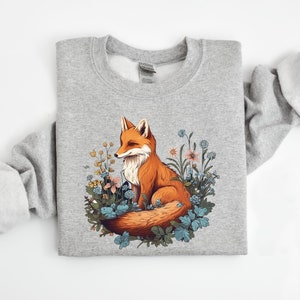 May include: A heather gray crewneck sweatshirt featuring a central graphic illustration of a red fox sitting amidst a cluster of blue, yellow, and orange wildflowers and green foliage. The fox has a white chest and a bushy tail curled around its body.