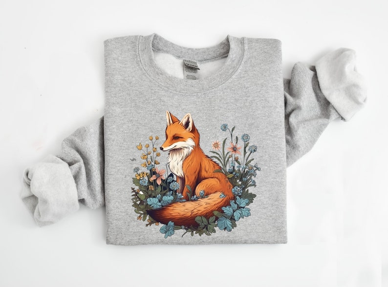 May include: A heather gray crewneck sweatshirt featuring a central graphic illustration of a red fox sitting amidst a cluster of blue, yellow, and orange wildflowers and green foliage. The fox has a white chest and a bushy tail curled around its body.