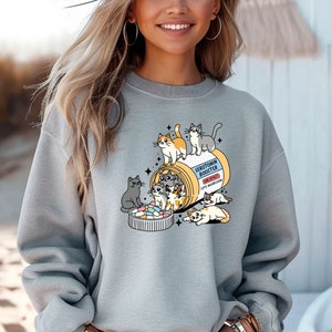 K&ouml;nnte beinhalten: Ein hellgraues Sweatshirt mit Rundhalsausschnitt und einer Grafik einer gelben Pillendose mit der Aufschrift Serotonin Booster sowie dem Text Side Effect Cute Aggression. Mehrere gezeichnete Katzen klettern auf, in und um die Dose.