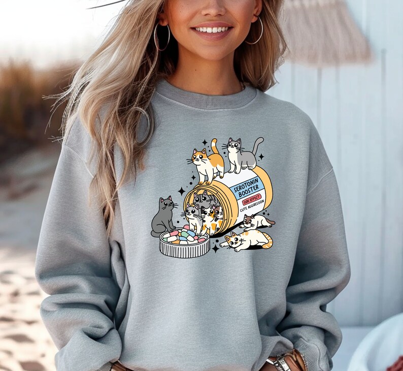 K&ouml;nnte beinhalten: Ein hellgraues Sweatshirt mit Rundhalsausschnitt und einer Grafik einer gelben Pillendose mit der Aufschrift Serotonin Booster sowie dem Text Side Effect Cute Aggression. Mehrere gezeichnete Katzen klettern auf, in und um die Dose.