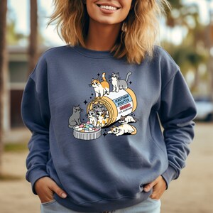 K&ouml;nnte beinhalten: Ein schieferblaues Sweatshirt mit Rundhalsausschnitt und einer Grafik einer gelben Pillendose mit der Aufschrift Serotonin Booster und dem Nebenwirkungshinweis Cute Aggression. Mehrere Cartoon-Katzen klettern auf und um die Dose.