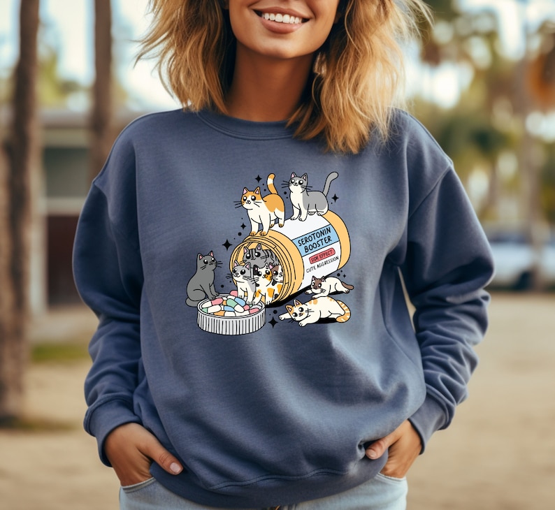 K&ouml;nnte beinhalten: Ein schieferblaues Sweatshirt mit Rundhalsausschnitt und einer Grafik einer gelben Pillendose mit der Aufschrift Serotonin Booster und dem Nebenwirkungshinweis Cute Aggression. Mehrere Cartoon-Katzen klettern auf und um die Dose.