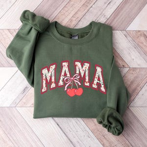 Może przedstawiać: Zielona bluza z napisem "MAMA" w kwiatowym wzorze i dwiema czerwonymi wiśniami z r&oacute;żową kokardką.