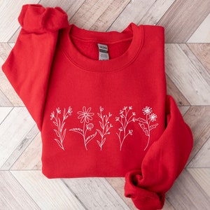 May include: A bright red long-sleeve crewneck sweatshirt featuring a white line art graphic of five distinct wildflowers arranged in a horizontal row across the center. The garment has a ribbed collar and cuffs with a Gildan brand label inside.