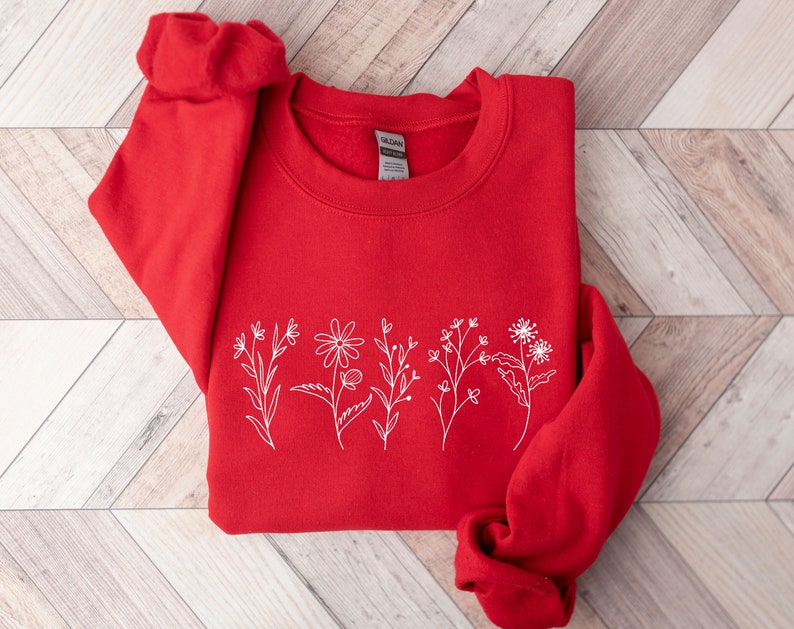 May include: A bright red long-sleeve crewneck sweatshirt featuring a white line art graphic of five distinct wildflowers arranged in a horizontal row across the center. The garment has a ribbed collar and cuffs with a Gildan brand label inside.