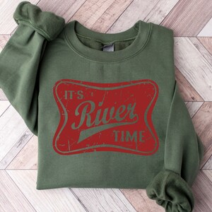 May include: An olive green crewneck sweatshirt featuring a distressed red rectangular graphic with rounded corners. The text "IT'S River TIME" is printed in olive green across the center of the red graphic, matching the color of the sweatshirt fabric.